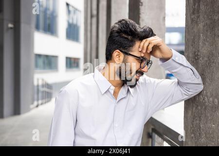Tired young Indian man standing near a building, leaning against a wall and holding his head with his hand, suffering from pain and pressure. Close-up photo. Stock Photo