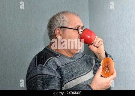 The picture shows an elderly man drinking from a cup and holding a pie in his other hand. Stock Photo