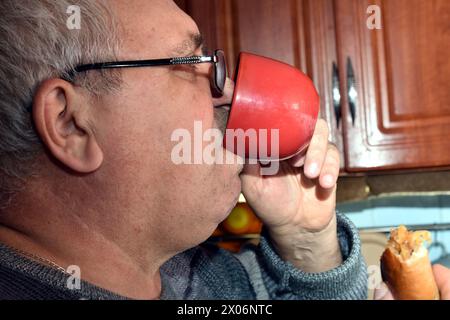 The picture shows an elderly man drinking from a cup and holding a pie in his other hand. Close-up. Stock Photo
