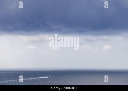 A small speedboat leaves a white wake across calm blue water beneath a broad bank of dark clouds, with a pale horizon separating sea and sky. Stock Photo
