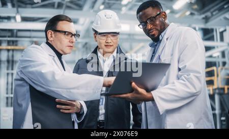 Team of Diverse Professional Heavy Industry Engineers Wearing Safety Uniform and Hard Hats Working on Laptop Computer. Technician and Workers Talking on a Meeting in a Factory Facility. Stock Photo