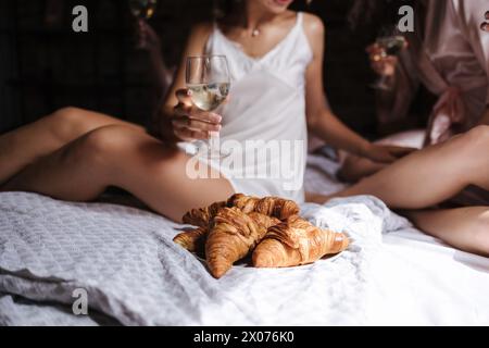 Close-up of four hands hold champagne with blueberry. Females sits on bed. Woman wears silk robe. Croissant on plate Stock Photo