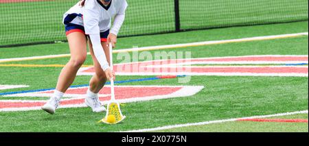 Front view of a girls lacrosse player scooping up the ball during a game on a green turf field. Stock Photo