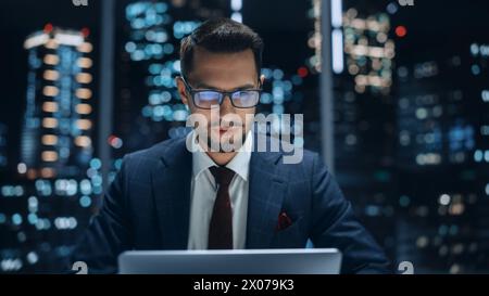 Portrait of a Successful Handsome Businessman Working on Laptop Computer in Big City Office Late in the Evening. Happy Competent Manager in Stylish Dark Blue Suit Doing Quarter Report. Stock Photo