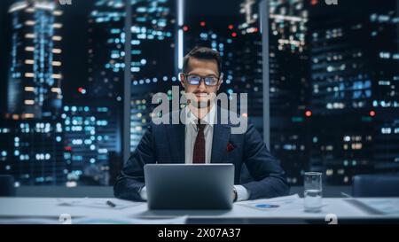 Portrait of a Successful Handsome Businessman Working on Laptop Computer in Big City Office Late in the Evening. Happy Competent Manager in Stylish Dark Blue Suit Smiles and Poses for Camera. Stock Photo
