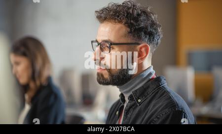 Portrait of a Handsome Concentrated Smart Male Student, Studying in Modern University. He Wears Glasses and Has a Beard and Short Curly Hair. Works on Computer in College. Stock Photo