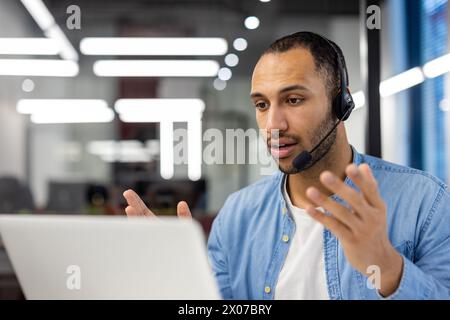 Serious young hispanic man in headset sitting at desk in office and talking on video call. Close-up photo. Stock Photo