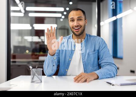 Cheerful Indian businessman in a casual blue shirt engaging in a video call from his modern office, greeting with a wave. Stock Photo