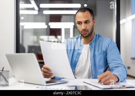 Focused Indian businessman reviewing documents while working remotely from a contemporary living room, exuding professionalism and dedication. Stock Photo