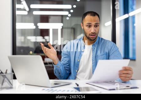 Surprised Indian man in a living room setting, dressed as a businessman, reacting to a paper document. Concept of unexpected news in home office environment. Stock Photo