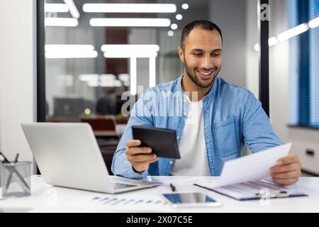 Focused Indian businessman in a modern office setting, working on his laptop while consulting documents on a desk. Stock Photo