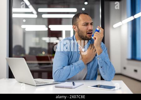 A man of Indian descent is in an office setting, using an asthma inhaler, showing signs of respiratory distress. Stock Photo