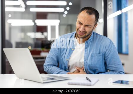 Indian businessman experiencing stomach pain while working on a laptop in a modern office setting, appearing distressed. Stock Photo