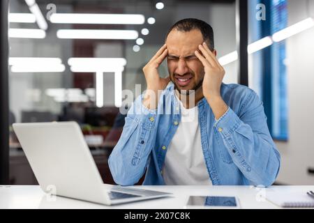 An Indian man feeling stressed and holding his head while working on a laptop in a modern office setting. Stock Photo