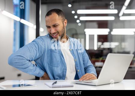 An Indian man in a living room setting appears to suffer from back pain while working on his laptop, portraying common workplace discomfort. Stock Photo
