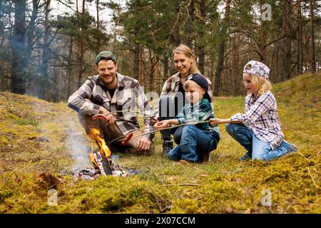 family with two children frying sausages over a bonfire while camping in forest. family time, nature adventure Stock Photo