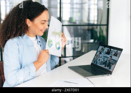 Financial online consultation. Brazilian businesswoman, ceo, team leader, sit at work dest in the office in front of a laptop, talks by online video conference with multiracial team, having brainstorm Stock Photo