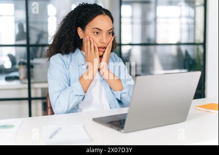 Confused brazilian or hispanic curly business woman, corporate financial manager, sit in modern office, puzzled looking at laptop screen, grabbed her cheeks with her hands, received unexpected news Stock Photo