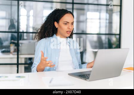 Unsatisfied brazilian or hispanic curly business woman, corporate financial manager, sit in modern office, puzzled looking at laptop screen, received unexpected news, feels at a loss Stock Photo