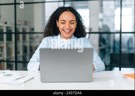 Working in the office. Happy successful brazilian or hispanic woman, company seo manager, sits in a modern office space at work desk with laptop, working on a project, develop strategy, smiles Stock Photo