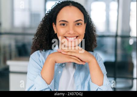 Close-up photo of a gorgeous successful latino or brazilian curly woman, company employee, software manager, sitting in the modern office space, looks at camera with friendly smile Stock Photo