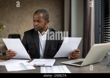 A man in a suit is holding two pieces of paper in his hands, looking at them with a frown on his face Stock Photo
