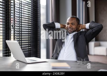 A man in a suit is sitting at a desk with a laptop and a notebook. He is relaxed and smiling, possibly taking a break from work Stock Photo