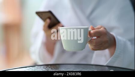 Cropped photo of a woman in a terry bathrobe holding mobile phone and cup Stock Photo