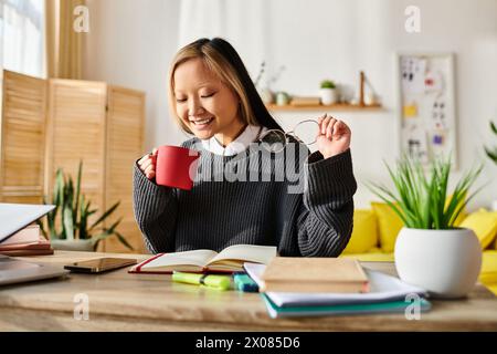 A young Asian woman sits at a table, deep in thought while enjoying a cup of coffee, surrounded by books and a laptop. Stock Photo