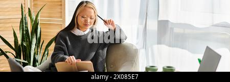 A young Asian woman sits in a chair, holding a book, surrounded by learning materials Stock Photo