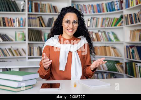 A young woman actively engaging with someone unseen, using expressive hand gestures, sitting in front of a bookshelf with books. Stock Photo