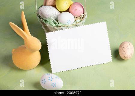 White blank note standing on pastel yellow green backdrop surrounded by decorative color Easter eggs and yellow bunny. Holiday decorations. Festive ca Stock Photo