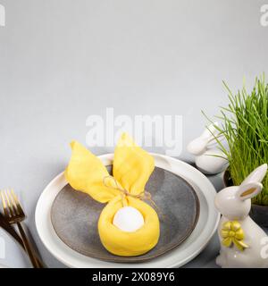 Plates, white egg in yellow napkin as bunny ears, fresh green grass, white ceramic bunnies, gold cutlery. Easter table setting composition on gray bac Stock Photo