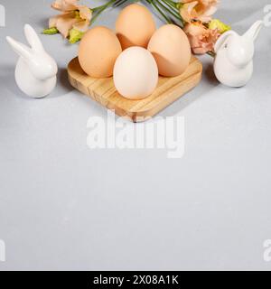Organic Easter card in trendy colors. Natural eggs in wooden egg stand, gentle peach Eustoma flowers, two white ceramic bunnies on gray background. Sq Stock Photo