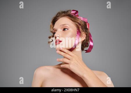 Young woman exudes classic beauty as she confidently poses with a pink bow on her head in a studio setting on a grey background. Stock Photo