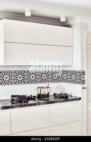 Modern kitchen design featuring white cabinetry, a patterned tile backsplash, and a stainless-steel pot on a gas stove Stock Photo
