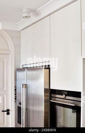 Sleek and stylish modern kitchen featuring high-end stainless steel refrigerator and built-in oven set against white cabinetry Stock Photo