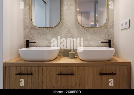 A contemporary bathroom vanity featuring two oval vessel sinks, matte black faucets, a textured accent wall, and a sleek mirror with a gold frame Stock Photo