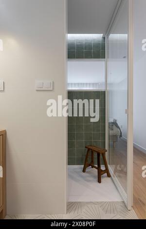 An elegant modern bathroom featuring a transparent glass shower door, green tiles, and a wooden stool Stock Photo