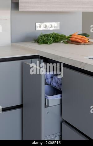 A sleek gray kitchen drawer is open to reveal neatly rolled linens, with fresh carrots and herbs on the countertop in the background Stock Photo