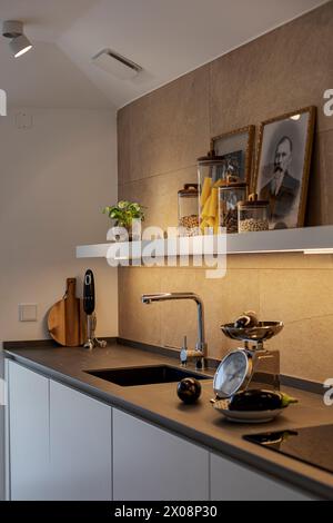 A stylish modern kitchen featuring a sleek black countertop, framed vintage portraits, and elegant storage jars with pasta and legumes Stock Photo