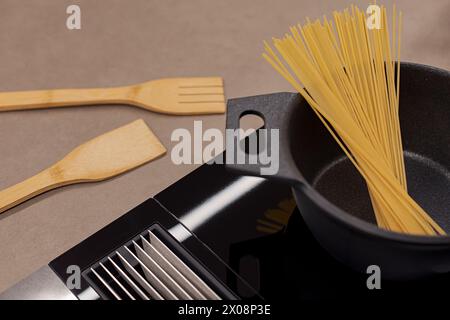 A cast iron pan sits on a modern induction cooktop, with raw spaghetti noodles and wooden utensils ready for cooking Stock Photo