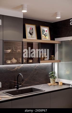 Modern kitchen interior featuring dark marble countertops, framed rooster art, and elegant cabinetry, with natural light filtering in Stock Photo