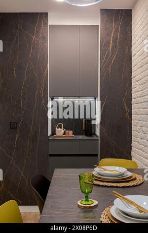 Elegantly designed kitchen space featuring black marble walls with distinctive veining, sleek cabinetry, and a well-lit dining area with colorful chai Stock Photo