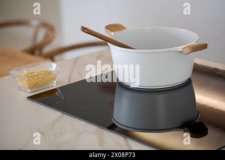 A white cooking pot rests on an induction cooktop with a wooden spoon, beside a bowl of raw pasta, in a stylish modern kitchen setting Stock Photo