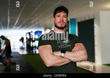 A determined young man stands with his arms crossed, wearing workout attire, in a modern gym setting. Stock Photo