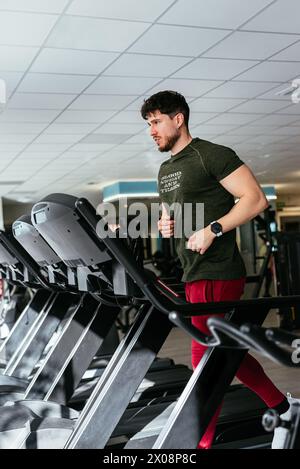 A concentrated young male wearing a green t-shirt and red shorts is running on a treadmill, staying fit in a modern gym setting. Stock Photo