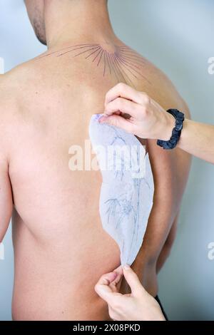 A cropped unrecognizable tattoo artist is carefully applying a transfer paper with a feather design onto the back of a male client in preparation for Stock Photo