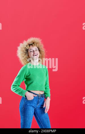 A joyful woman with a beaming smile and curly hair poses playfully, wearing a cozy green sweater and classic blue jeans on a bright red background Stock Photo