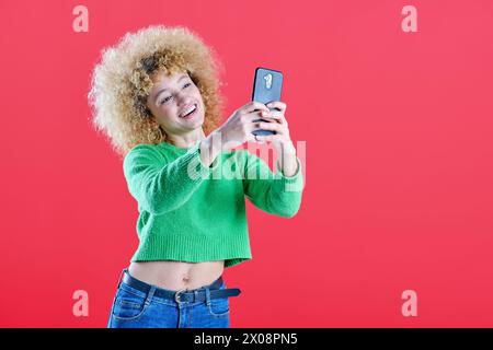 An exuberant curly-haired woman takes a selfie with a joyful smile, wearing a vibrant green sweater against a vivid red background Stock Photo